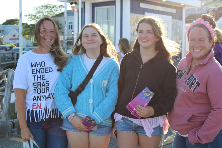 Four smiling women outdoors, two holding pamphlets, one wearing a blue cardigan.