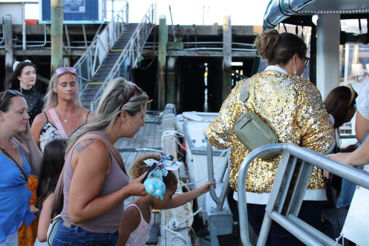 People boarding a boat, woman in gold sequin jacket, docks and stairs in the background.