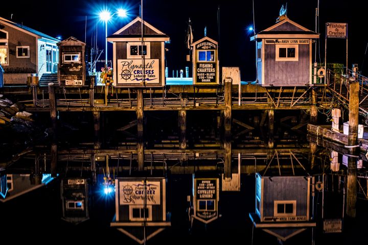 Night scene of wooden boat rental shacks reflecting in calm water, with bright lights illuminating the area.