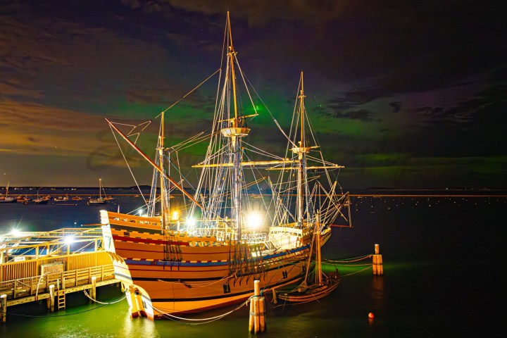 Illuminated tall ship docked at night with a dark, cloudy sky and numerous smaller boats in the background.
