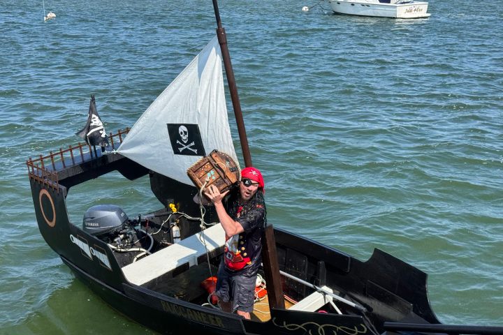 Person dressed as pirate on small boat with treasure chest, surrounded by water and other boats.