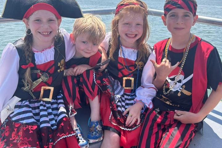 Four children in pirate costumes posing on a boat with a scenic view of water and sky.