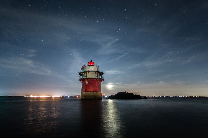 Lighthouse under starry sky, surrounded by calm water at night.