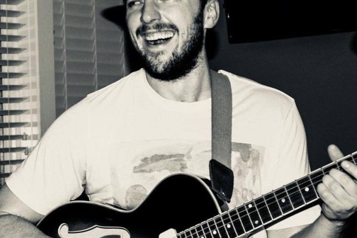 Smiling man playing guitar indoors, wearing a hat.