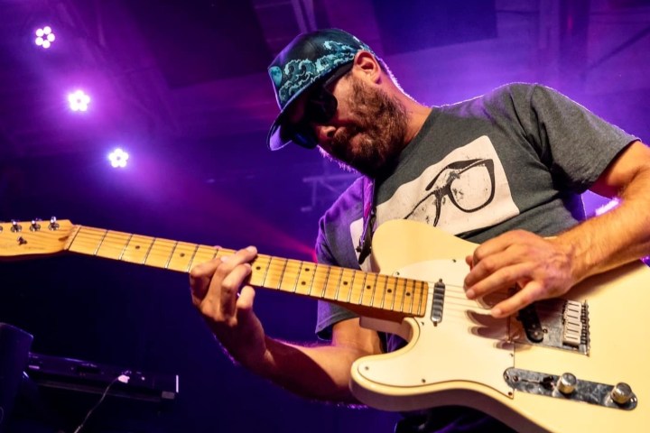 Man playing electric guitar on stage under purple lighting.