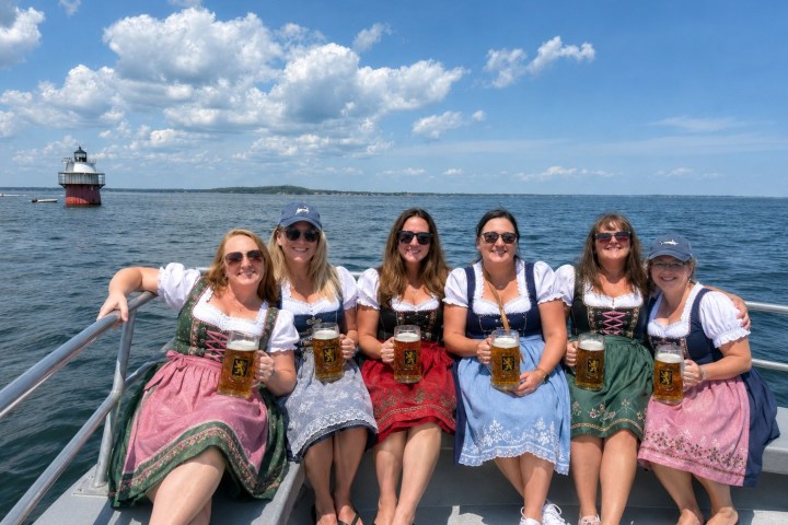 Six women in traditional dresses holding beer mugs on a boat near a lighthouse.